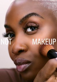 En makeupborste med ett mjukt borsthuvud appliceras på en jämn, djupt brun hudton med en guldsnäsring; fet text säger "INTE MAKEUP."