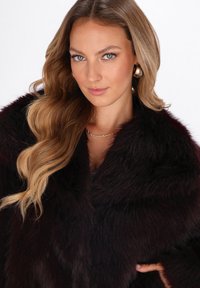 Žena s dugom valovitom platinastom kosom koja nosi zlatne kružne naušnice, delikatnu ogrlicu i tamnosmeđi krzneni kaput na bijeloj pozadini.