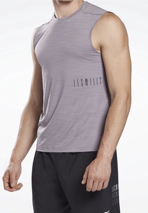 Mann trägt ärmelloses hellviolettes Sportshirt und schwarze Shorts mit "Les Mills" Logo auf beiden Kleidungsstücken, steht vor weißem Hintergrund.