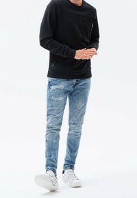 Sudadera negra con bolsillo frontal, jeans desgastados de color azul claro y zapatillas blancas con un diseño estampado, de pie sobre un fondo simple.