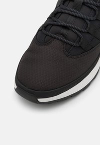 Shoe atlético preto com parte superior em malha texturizada, apresentando detalhes padronizados e sola de borracha grossa com faixa branca ao longo da borda.