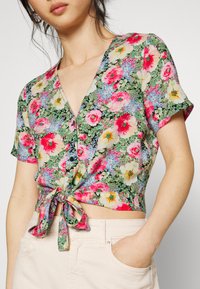 Top envolvente floral com um padrão vibrante de flores rosa e brancas sobre um fundo verde, com um laço na cintura e mangas curtas.