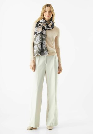 Woman wearing beige sweater, white wide-leg pants, beige shoes, and a large black, gray, and beige floral-patterned scarf.