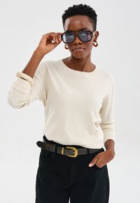 Lekka, kremowa sweter z prążkowanymi mankietami, w połączeniu z czarnymi spodniami o wysokim stanie i brązowym paskiem z złotą klamrą. Duże, ciemne okulary przeciwsłoneczne.