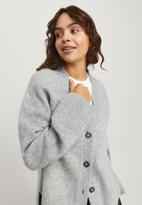 Cardigan en laine grise avec un décolleté en V, des poignets côtelés et cinq boutons ronds. Présente des fentes sur les côtés pour plus de confort et une coupe décontractée.