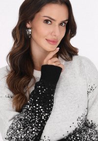 Pulover gri cu accente din paiete negre pe mâneci, manșete cu nervuri și textură moale. Modelul pozează cu mâna sub bărbie.