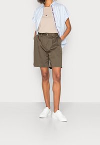 Osoba oblečená v světle modré pruhované košili s krátkým rukávem přes béžové tričko Tommy Hilfiger, olivově zelené šortky s páskem a bílé tenisky.