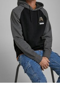 Felpa con cappuccio nera e grigia con etichetta del logo, maniche raglan, cappuccio con coulisse e vestibilità comoda. Indossata con pantaloni cargo blu.