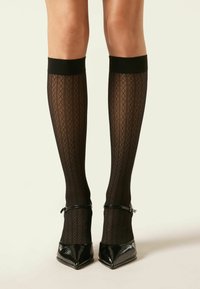 Calcetines negros hasta la rodilla con un patrón de diamantes y una textura suave, rematados con una banda negra sólida; combinados con tacones de charol negros de punta.