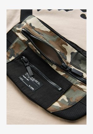 Tarnfarbenes, mit Reißverschluss versehenes Fach mit schwarzen Akzenten, featuring eine strukturierte Oberfläche und ein Etikett. Der beige Stoffhintergrund ist sichtbar.