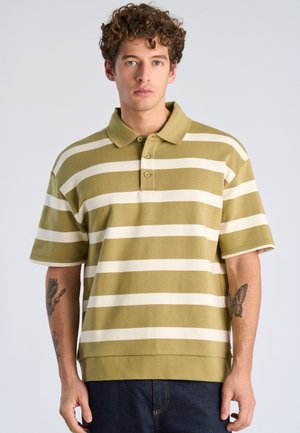 Jeune homme aux cheveux bouclés portant un polo à manches courtes rayé olive et blanc, montrant des tatouages d'aigle et de papillon sur les avant-bras.