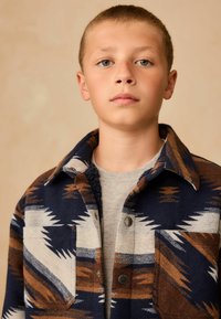 Réteges kabát kék, barna és fehér geometrikus mintával, texturált anyagból, inggallérral és fémből készült gombokkal.