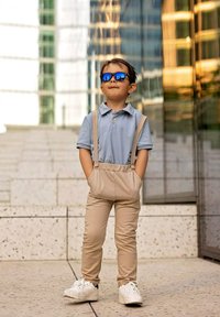 Jasnoniebieska koszulka polo, beżowe spodnie z szelkami, białe trampki. Dziecko w niebieskich okularach przeciwsłonecznych, stojące z rękami w kieszeniach, na tle odbijającej powierzchni.