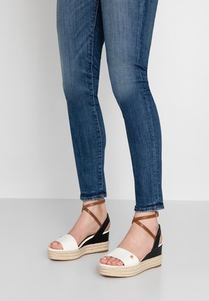 Pieds portant un jean skinny bleu et des sandales compensées beige avec des talons noirs et des brides marron à la cheville sur un sol gris.