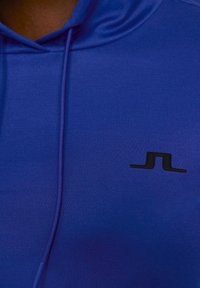 Blå hoodie i texturerad tyg med en svart logotyp på framsidan. Designen inkluderar en justerbar huva med dragsko.