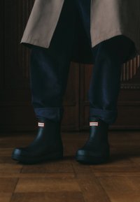 Schwarze Gummistiefel bis zum Knöchel mit rotem "HUNTER"-Label, runder Zehenform und niedrigem, strukturiertem Sohlen, kombiniert mit marineblauen Cordhosen.