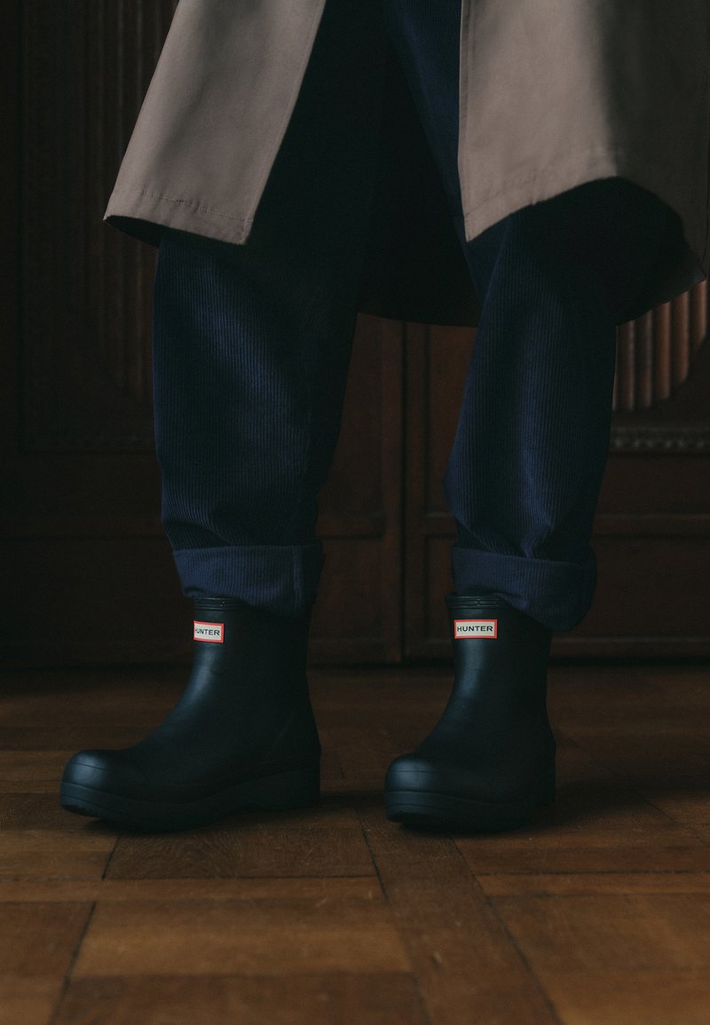 Schwarze Gummistiefel bis zum Knöchel mit rotem "HUNTER"-Label, runder Zehenform und niedrigem, strukturiertem Sohlen, kombiniert mit marineblauen Cordhosen.