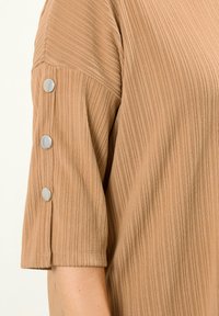 Brauner, gerippter Stofftop mit kurzen Ärmeln und silbernen Druckknöpfen an den Bündchen, betont Textur und klare Linien.