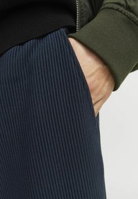 Pantalone od tamnoplave rebraste tkanine s vidljivim bočnim džepom, uparen s tamnozelenim rukavima s manžetama, ističući kontrastne teksture i boje.