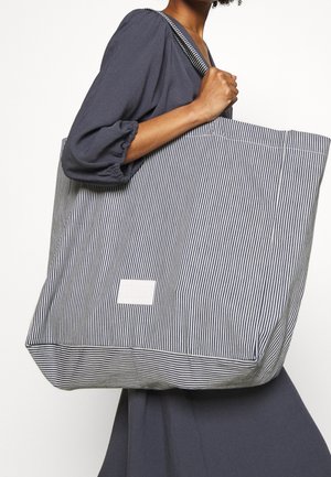 Sac en tissu rayé avec une base carrée, doté d'une étiquette blanche, de poignées robustes et d'une finition texturée dans des couleurs bleu marine et blanche.