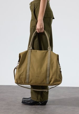 Persoon in olijfgroene broek en zwarte schoenen houdt een grote olijfgroene canvas tas met bovenhandvatten en schouderriem vast.