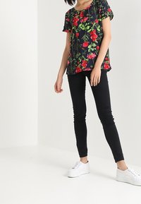 Bluse mit Floralprint, kurzen Ärmeln, rundem Halsausschnitt und dunklem Hintergrund; kombiniert mit schwarzen Skinny Jeans und weißen Sneakers.