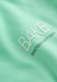 Muntgroene stof met een textuur en een verhoogd wit logo "BAKER" en daaronder in grijs "NINETEEN EIGHTY EIGHT".