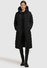 Femeie îmbrăcată într-un palton lung, negru, capitonat, cu glugă și închidere cu fermoar, purtând colanți negri și botine negre, stând pe un fundal simplu.