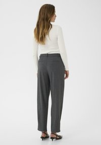 Kvinde med langt hår iført en hvid langærmet bluse, mørkegrå skræddersyede bukser og sorte lave hæle slingback sko, stående med ryggen til.