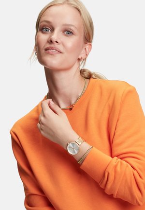 Montre en or avec un cadran blanc et un bracelet en métal, portée à la main. Affiche un sweatshirt orange avec un col rond et des manches longues.