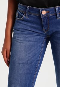 Jeans de denim azul oscuro con un diseño ajustado, estilo de cinco bolsillos y un detalle de botón de cobre en la cintura con costuras contrastantes.