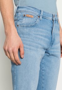 Jeans de denim azul claro com um corte reto, bolsos frontais e um rótulo de imitação de pele da Wrangler na cintura. Textura suave.