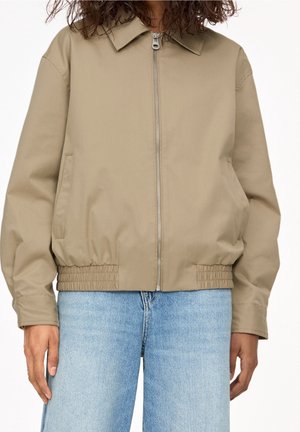 Veste beige zippée avec col et taille élastique portée avec un jean bleu clair sur une personne aux cheveux bouclés.