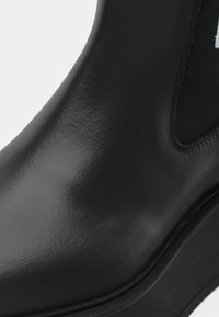 Bottine en cuir noir avec panneau latéral en élastique et semelle épaisse, montrant une surface lisse texturée et des détails de couture.