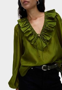 Zelená saténová blůza s hlubokým výstřihem do V, volánkovou lemovkou a zavazovacími detaily u krku; dlouhé rukávy; doplněno černým pásem a tmavými džíny.