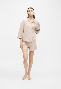 Jeune femme debout pieds nus portant une chemise beige à carreaux ample et un short assorti sur fond blanc uni.