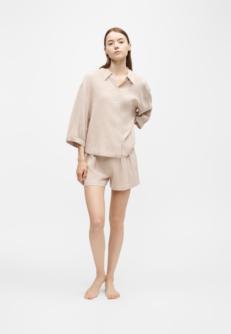 Jeune femme debout pieds nus portant une chemise beige à carreaux ample et un short assorti sur fond blanc uni.