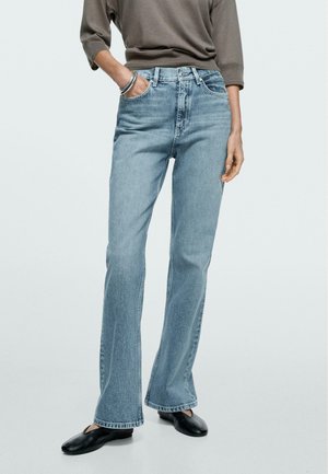 Kvinde iført lyseblå, højtaljede, straight-leg jeans, brun langærmet top, sorte flade sko og sølvfarvede armringe med den ene hånd i lommen.