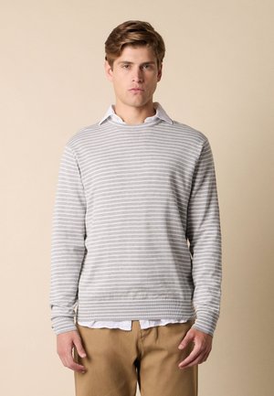 Jonge man met kort lichtbruin haar, die een grijs-wit gestreepte trui draagt over een wit overhemd en beige broek, staand tegen een beige achtergrond.