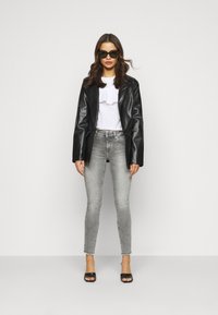 Casaco de pele preto, camisa branca com pregas, jeans cinzentos justos e sandálias pretas de salto. Modelo usando óculos de sol pretos, em pé.