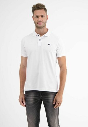 Man wearing a white short-sleeve polo shirt with black buttons and dark washed jeans standing against a white background.