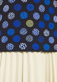 Mehrfarbiges Kleid mit einem marineblauen Oberteil, das verschiedene blaue und goldene Kreise zeigt, und einem hellbeigen, gefältelten Rock. Weicher Stoff mit auffälligen Mustern.