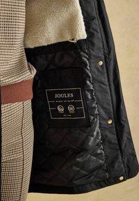Schwarze gesteppte Jacke mit flauschigem cremefarbenem Futter. Verfügt über ein Joules-Schild und Druckknöpfe. Das Innere enthält eine Tasche mit glatter Textur.