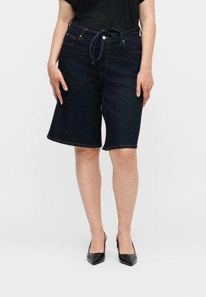 Femme portant un short en jean bleu foncé mi-long avec une ceinture nouée et des talons noirs à bout pointu sur un fond uni.