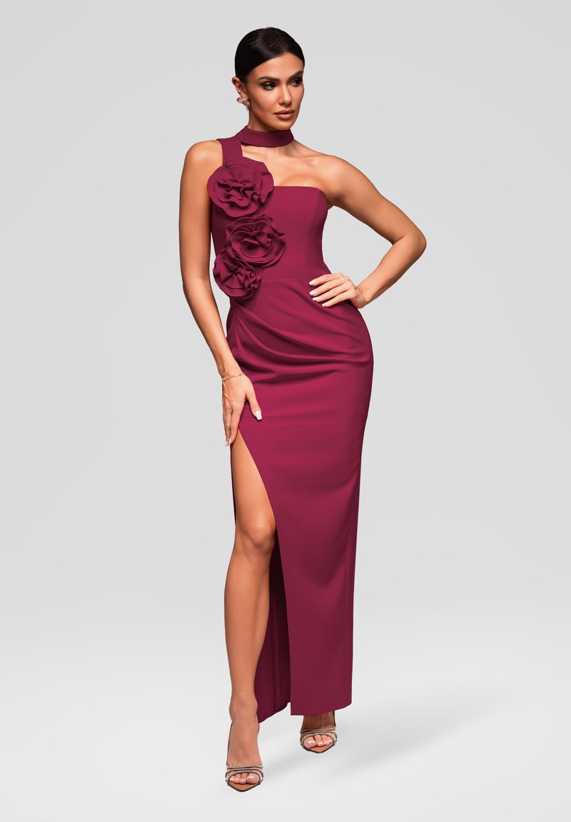 Femme posant dans une robe bordeaux sans manches avec une fente latérale et de grandes appliques florales sur le corsage, portant des talons à brides.