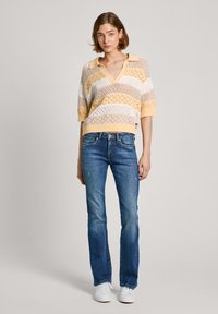 Stickad tröja med V-ringning och korta ärmar, med gula, vita och beige ränder; kombinerat med blå utsvängda jeans och vita sneakers.