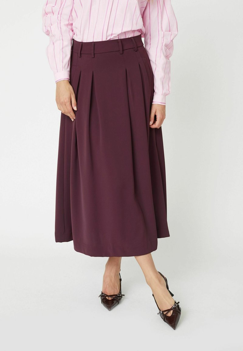 Una falda midi fluida de tejido burdeos oscuro, con pliegues y una cinturilla alta, combinada con zapatos negros de punta.