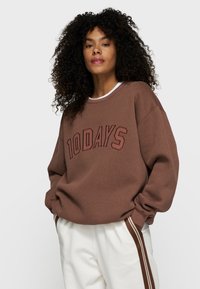 Brauner Pullover mit "10 DAYS" in dunklerem Braun geprägt. Besitzt einen runden Ausschnitt und einen oversized Fit, kombiniert mit hellen Hosen.