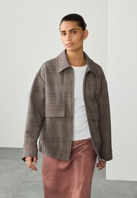 Giacca a quadri marrone con scollo con colletto, grande tasca frontale e chiusura con cerniera, abbinata a una maglia bianca e a una gonna rosa setosa.