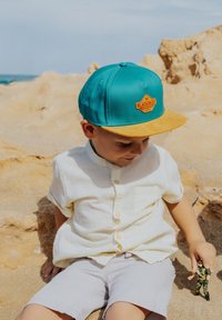 Gorra snapback turquesa con visera amarilla, que presenta un parche de logo naranja. El niño lleva una camisa ligera de botones y pantalones cortos beige.